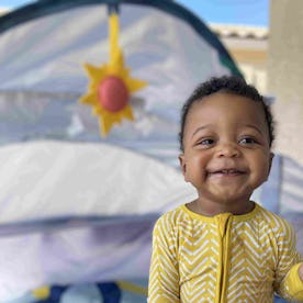 A joyful toddler in yellow pajamas holding a "My 1st ABC" book in front of a play tent with a sun de...