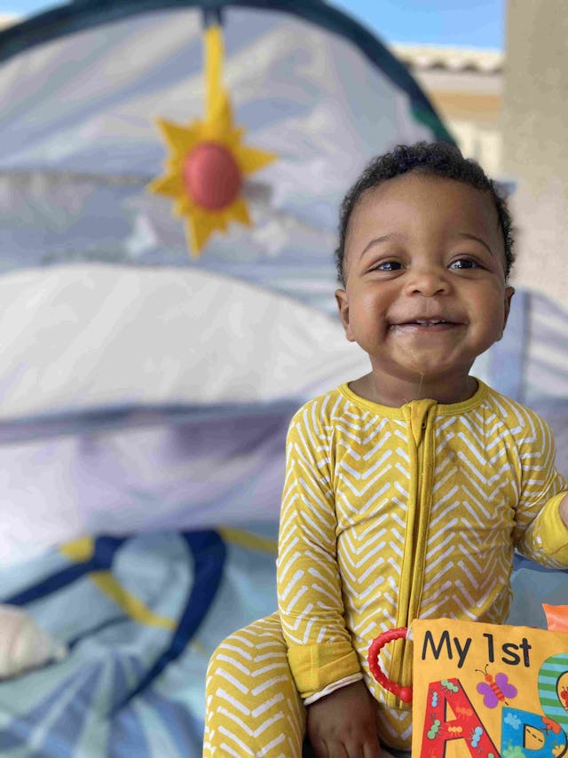 A joyful toddler in yellow pajamas holding a "My 1st ABC" book in front of a play tent with a sun de...