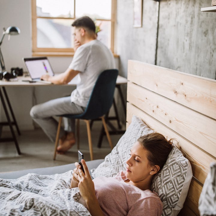 A woman laying on a bed checking her phone while a man is sitting at his desk with a laptop