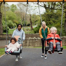 Two mom friends push their kids on the swings at the park.