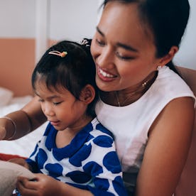 Asian mother and her little girl sitting comfortably on the bed at their home, focused on reading a ...