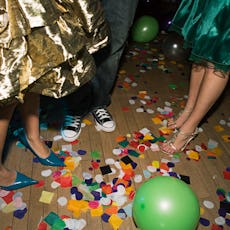 Two girls wearing dresses beside a boy in converse at a party with balloons on the floor around them...