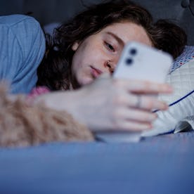 An Exhausted Caucasian brunette woman wakes up with a smartphone in hand after a restless night in b...