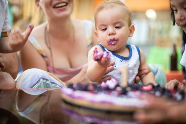 Toddler with cake on face at birthday party, surrounded by laughing family members.