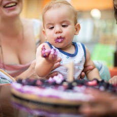 Toddler with cake on face at birthday party, surrounded by laughing family members.