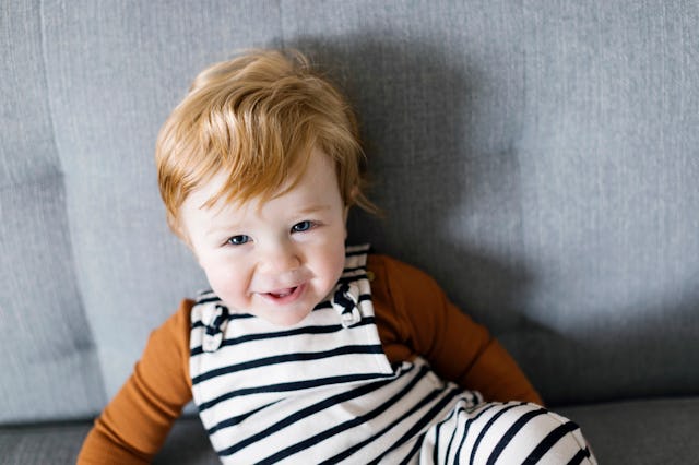 Toddler with red hair smiling, wearing striped overalls and an orange shirt, sitting on a grey sofa.