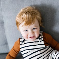 Toddler with red hair smiling, wearing striped overalls and an orange shirt, sitting on a grey sofa.