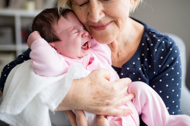 Elderly woman gently cradling and smiling at a crying newborn baby dressed in pink.