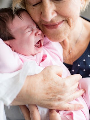 Elderly woman gently cradling and smiling at a crying newborn baby dressed in pink.