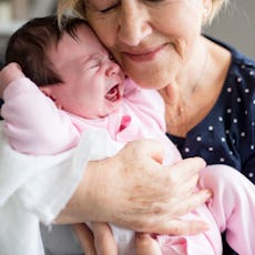 Elderly woman gently cradling and smiling at a crying newborn baby dressed in pink.