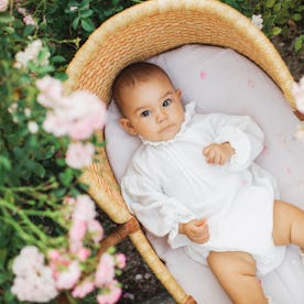 A baby in a vintage outfit lies in a bassinet outdoors.