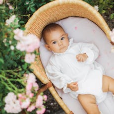 A baby in a vintage outfit lies in a bassinet outdoors.