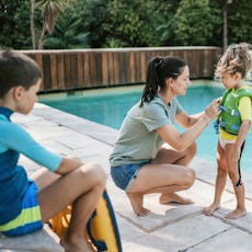 A mom snaps a life jacket on her daughter by the pool.