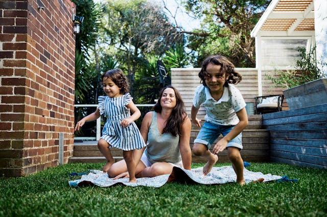 A mom sits on a blanket in the yard as her kids play.