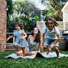 A mom sits on a blanket in the yard as her kids play.