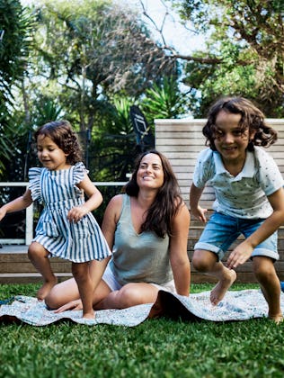 A mom sits on a blanket in the yard as her kids play.