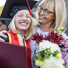 A joyful graduate in a cap and gown hugging an older woman while holding flowers at a graduation cer...