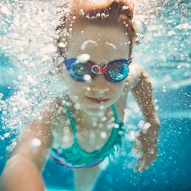 Young girl with goggles swimming underwater in a pool, surrounded by bubbles, looking at the camera.