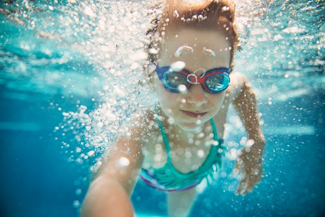 Young girl with goggles swimming underwater in a pool, surrounded by bubbles, looking at the camera.