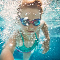 Young girl with goggles swimming underwater in a pool, surrounded by bubbles, looking at the camera.