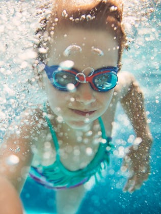 Young girl with goggles swimming underwater in a pool, surrounded by bubbles, looking at the camera.