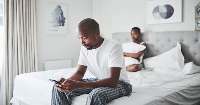 Husband sitting on the bottom side of the bed, looking at his phone. Wife is on the other side of th...