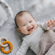 Portrait of a lovely Asian baby girl smiling sweetly while lying on the crib. She is raising her han...