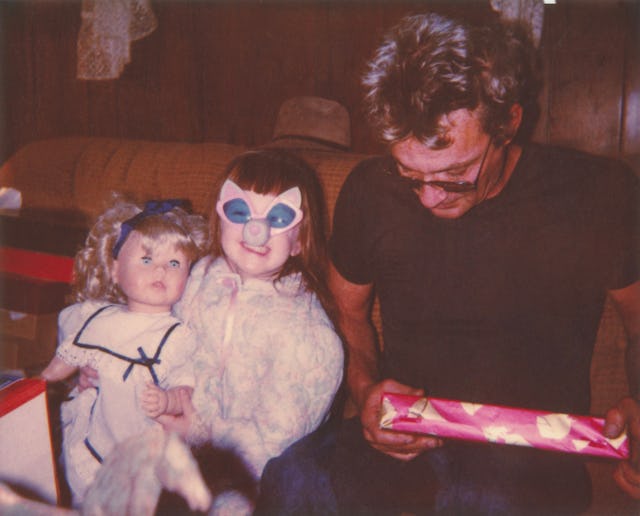 A man holding a gift sits beside his daughter on a couch; the daughter holds a doll and wears blue c...