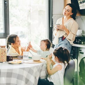 Tired mother drinks coffee while her tree daughter has breakfast at home.