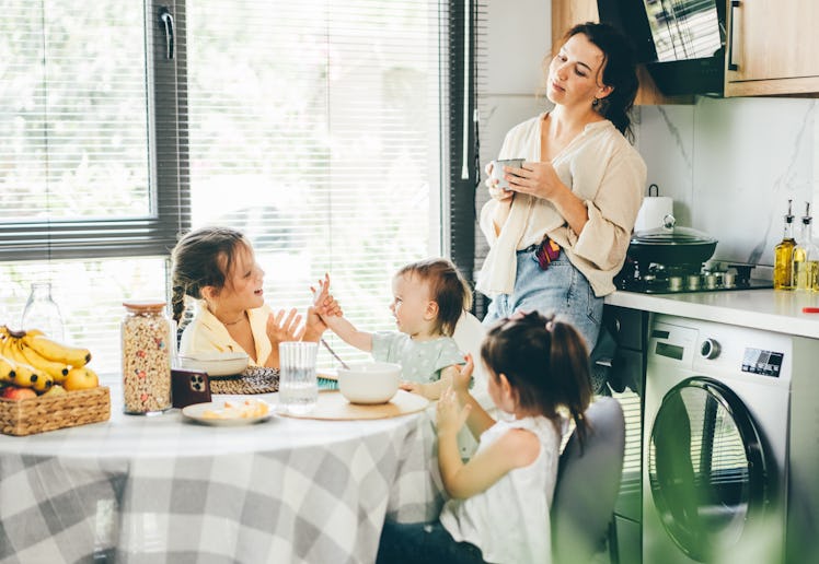 Tired mother drinks coffee while her tree daughter has breakfast at home.