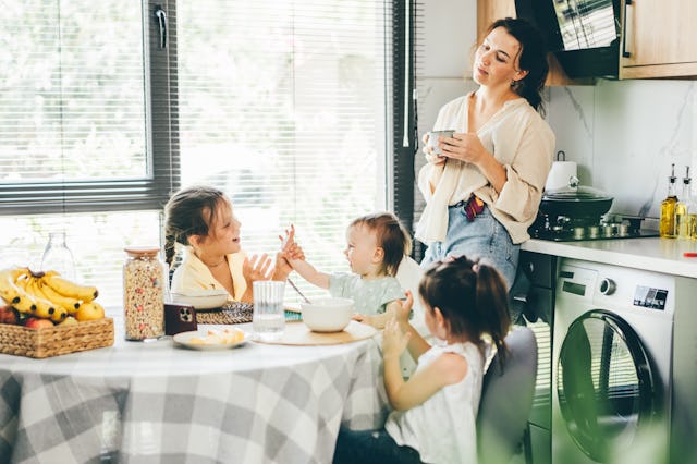 Tired mother drinks coffee while her tree daughter has breakfast at home.